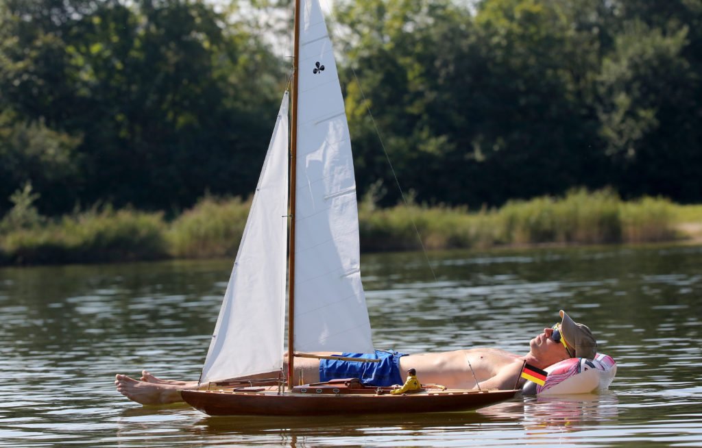 Un homme sur un matelas pneumatique prend un bain de soleil sur un lac tandis qu'un modèle réduit de bateau passe à côté de lui.